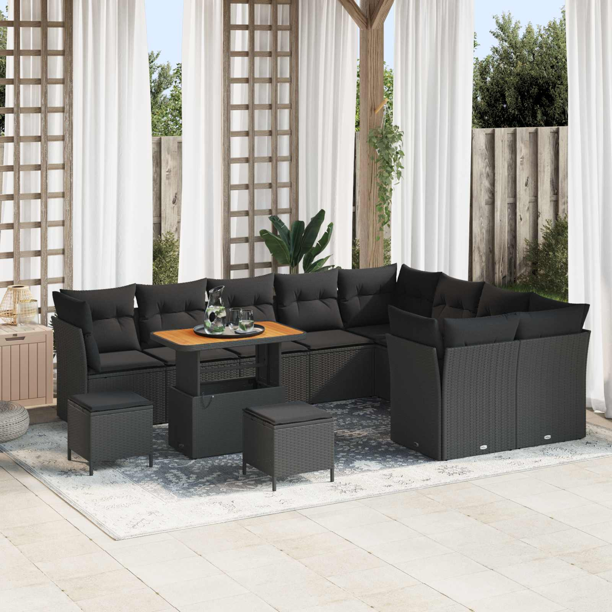 Outdoor-Schwarz-Wicker-Sofagarnitur mit Kissen, Tisch und Hockern auf einem Teppich, mit weißen Vorhängen und Holzgitter im Hintergrund.