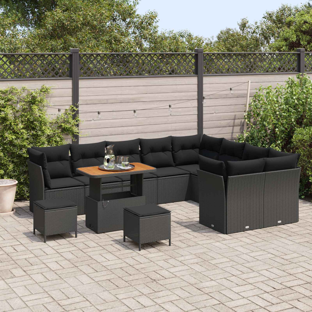 Garten-Terrassenset mit schwarzen Kissen, einem Holztisch und passenden Ottomanen. Auf einer gepflasterten Terrasse mit Zaunhintergrund.