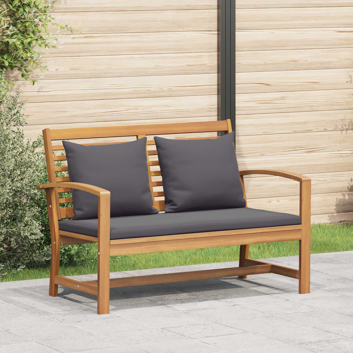Panca in legno con cuscini grigi. Lo sfondo mostra una staccionata di legno, verde e un patio pavimentato.
