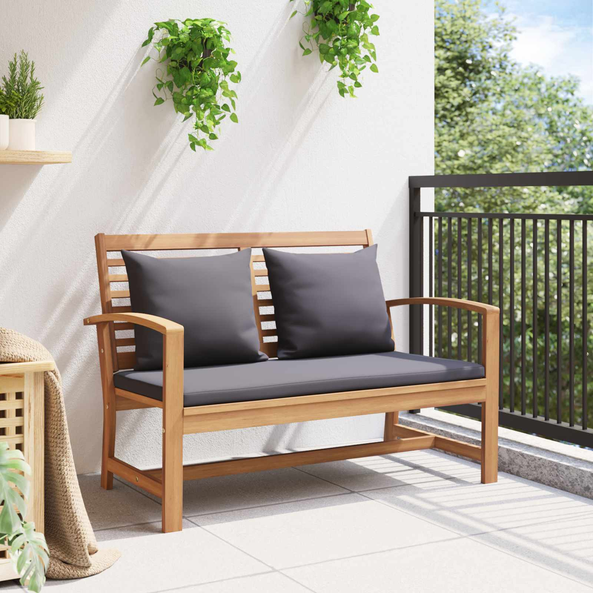 Panca in legno con cuscini grigi su un balcone. Piante verdi appese al muro bianco. Il balcone ha una ringhiera nera e vista sugli alberi.