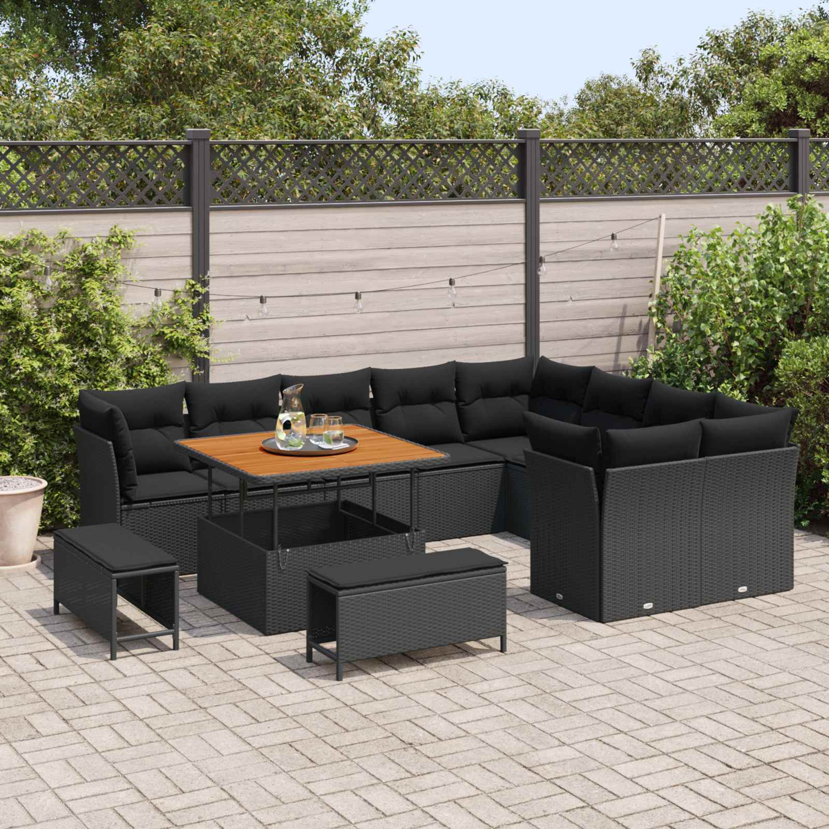 Schwarzes Outdoor-Rattan-Möbelset mit Tisch, Bänken und Ecksofa auf einer Terrasse mit Holzzaun.