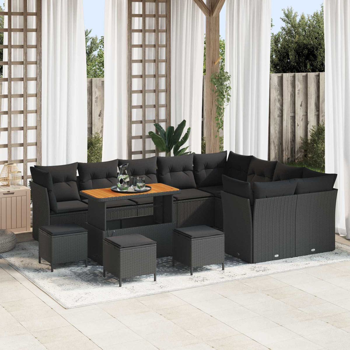 Outdoor-Sofa-Set aus schwarzem Korbgeflecht mit Kissen, Tisch und Hockern. Weiße Vorhänge und Blätter im Hintergrund.