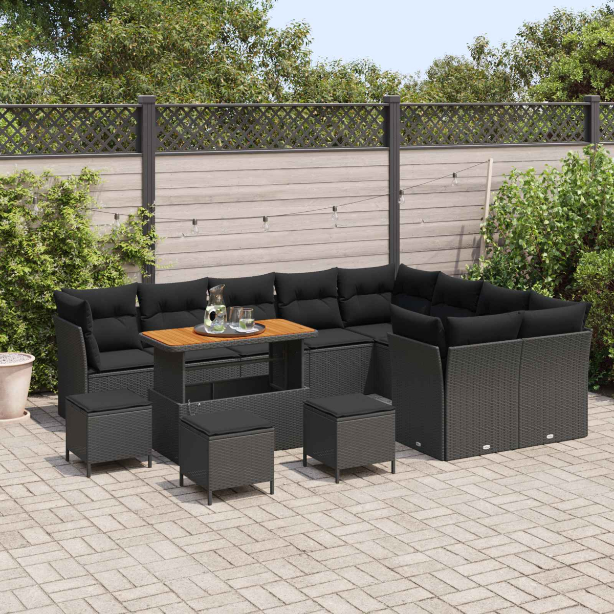 Terrassenset mit schwarzen Kissen, Holztisch und grau gewebten Möbeln. Auf einer gepflasterten Terrasse mit Holzzaun und Pflanzen.