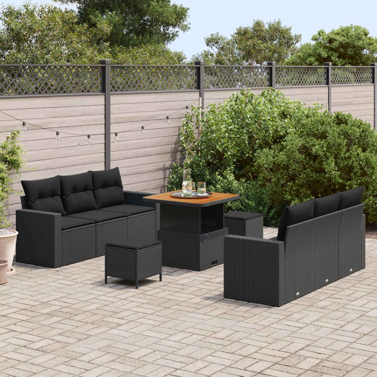 Juego de patio al aire libre con muebles de mimbre negro, que incluyen un sofá, sillas, otomanas y una mesa en un patio pavimentado con vegetación y una valla.