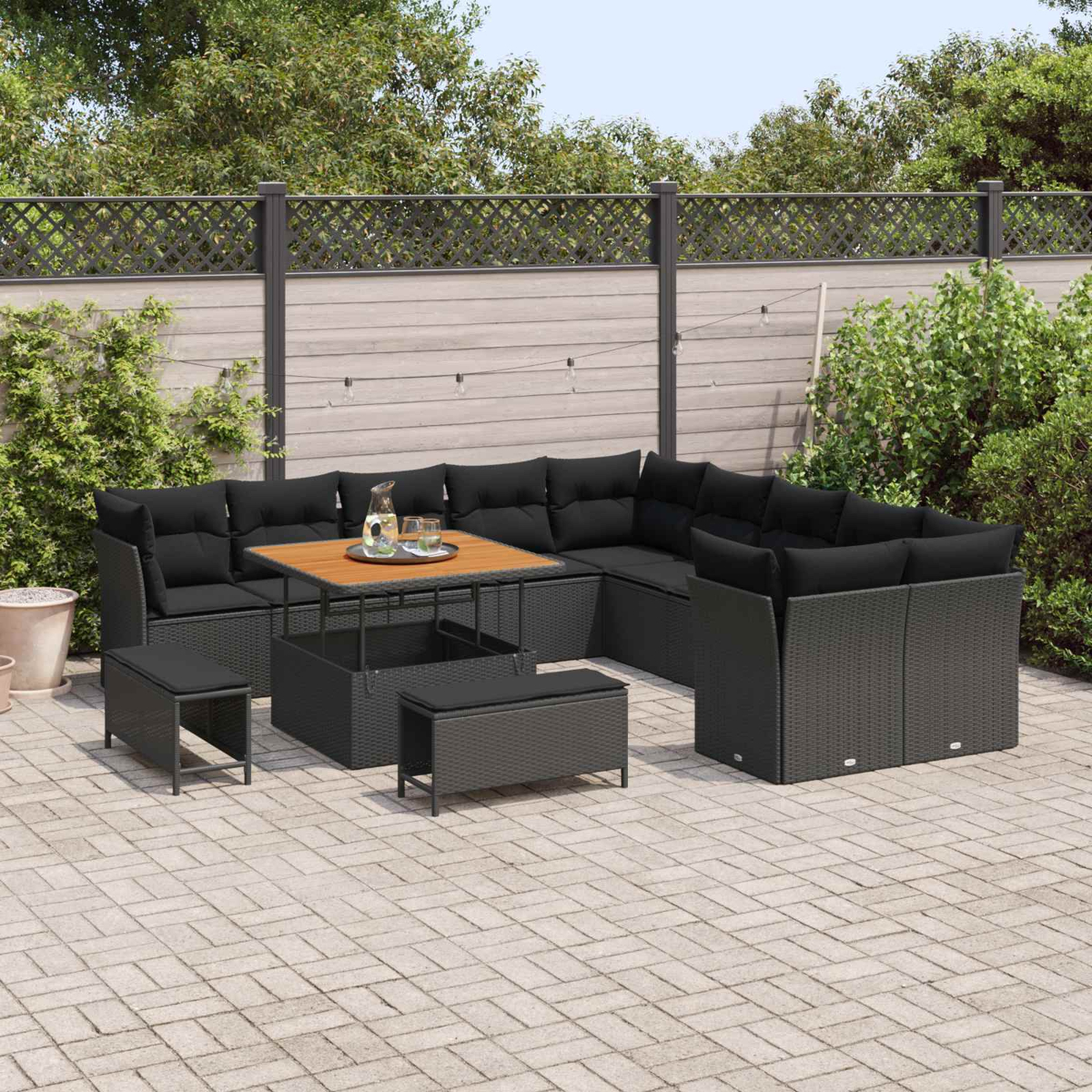 Outdoor-Möbelset aus schwarzem Korbgeflecht mit Kissen, Holztisch und kleinen Bänken auf einer Steinterrasse, mit einem Zaun im Hintergrund.