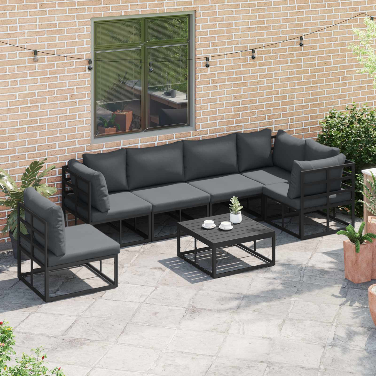 Set patio esterno con divano componibile, sedia e tavolino. Cuscini grigi, telai neri, su un patio in pietra con muro di mattoni.