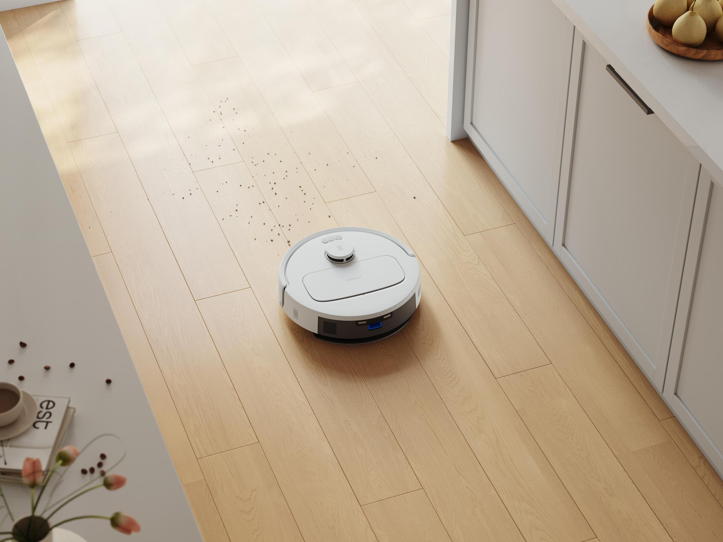 Ein weißer Roboterstaubsauger auf einem Holzboden mit verstreuten Trümmern. Küchenschrank und Tisch mit Blumen.