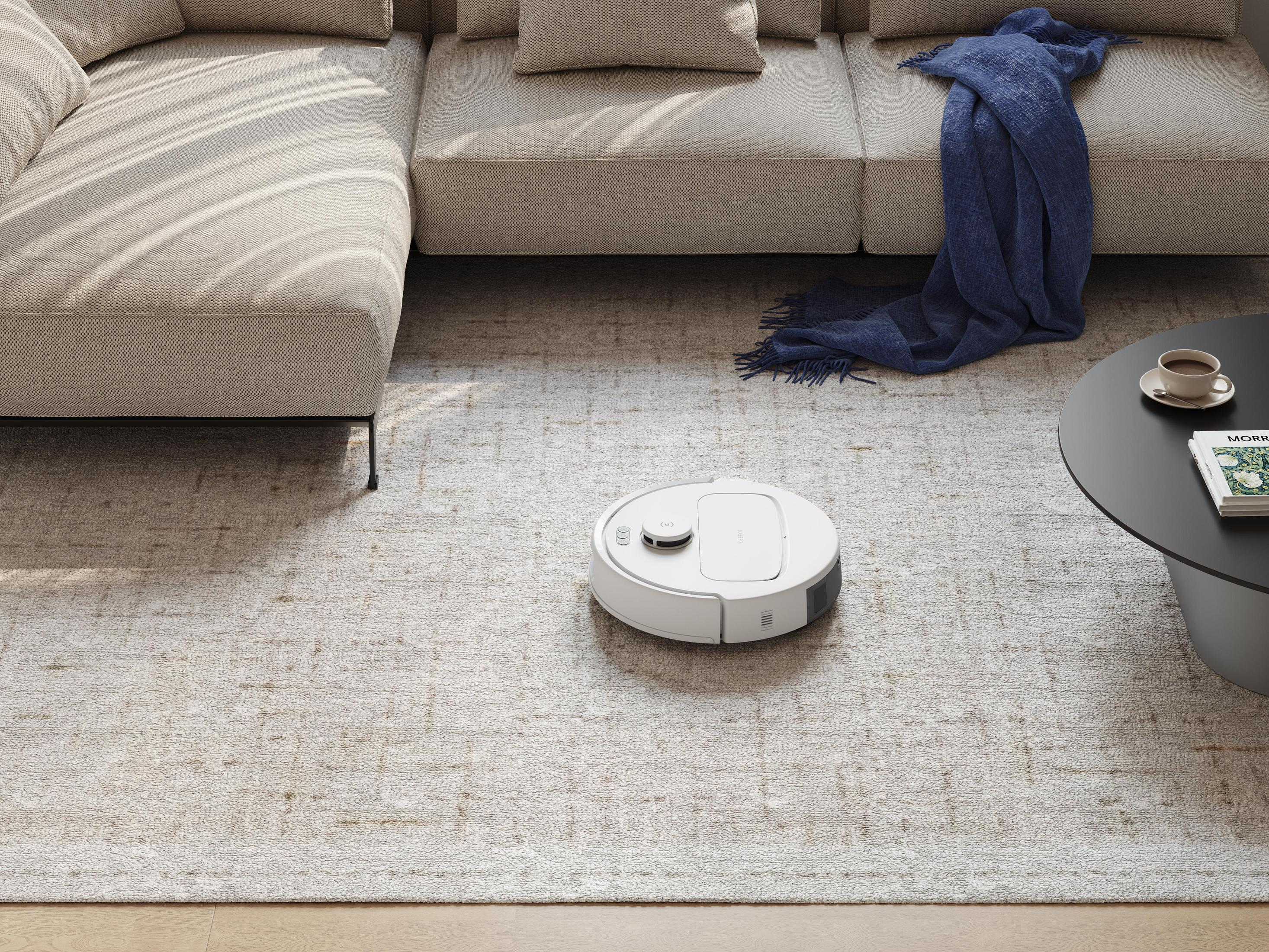 Ein weißer Roboterstaubsauger auf einem gemusterten Teppich in der Nähe eines Sofas und eines Couchtisches.