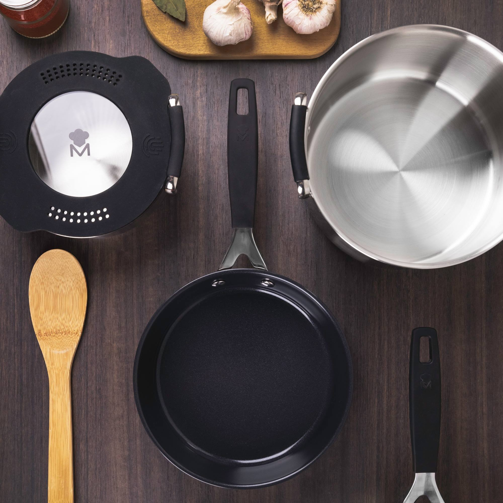 Vista superior de utensilios de cocina sobre superficie de madera: sartén, olla, tapa, espátula, cuchara, ajo y condimentos.