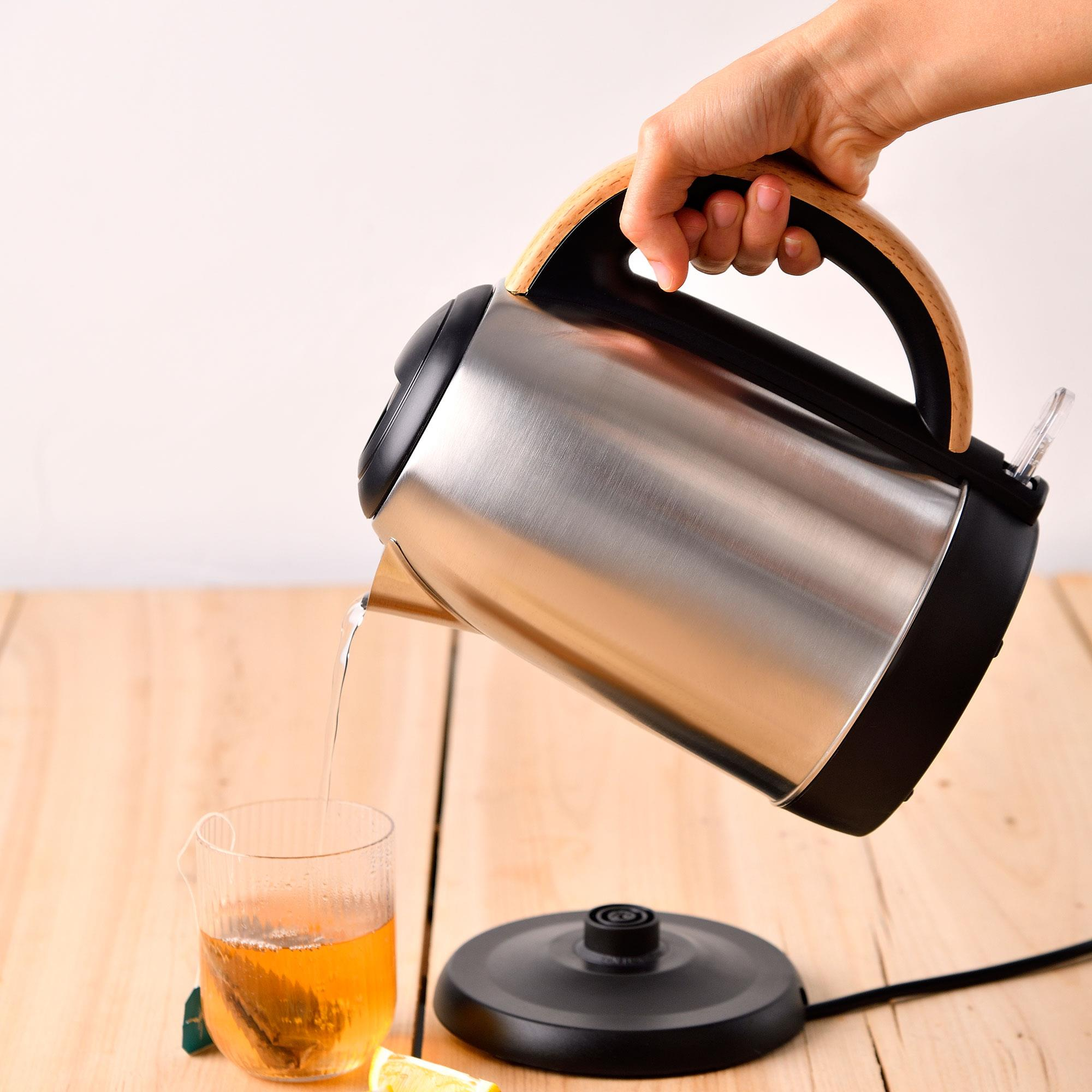 Una mano vierte agua de una tetera en un vaso de té. La tetera es de acero inoxidable con asa de madera.