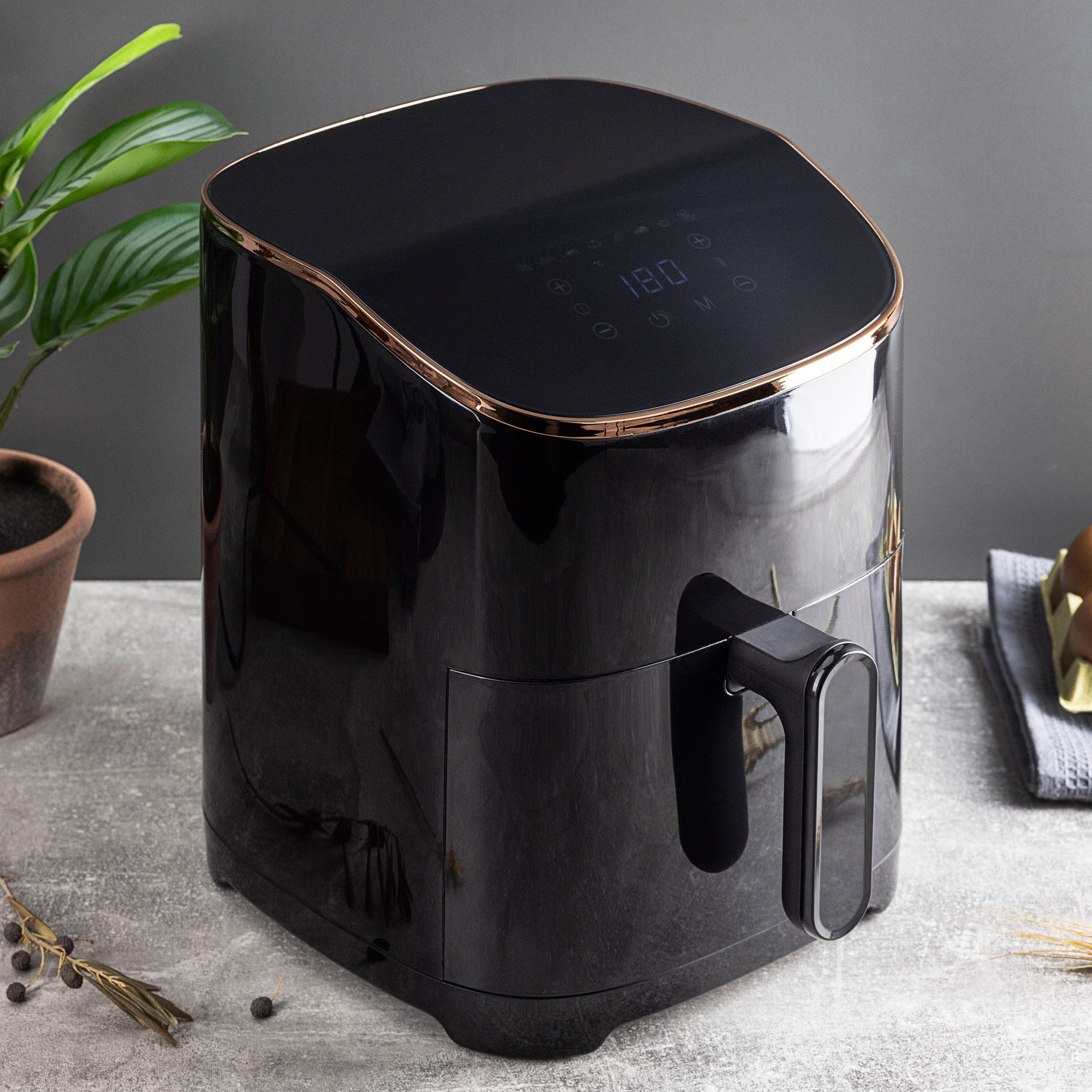 Friggitrice ad aria nera con finiture dorate su un tavolo. Pianta e ingredienti sullo sfondo.