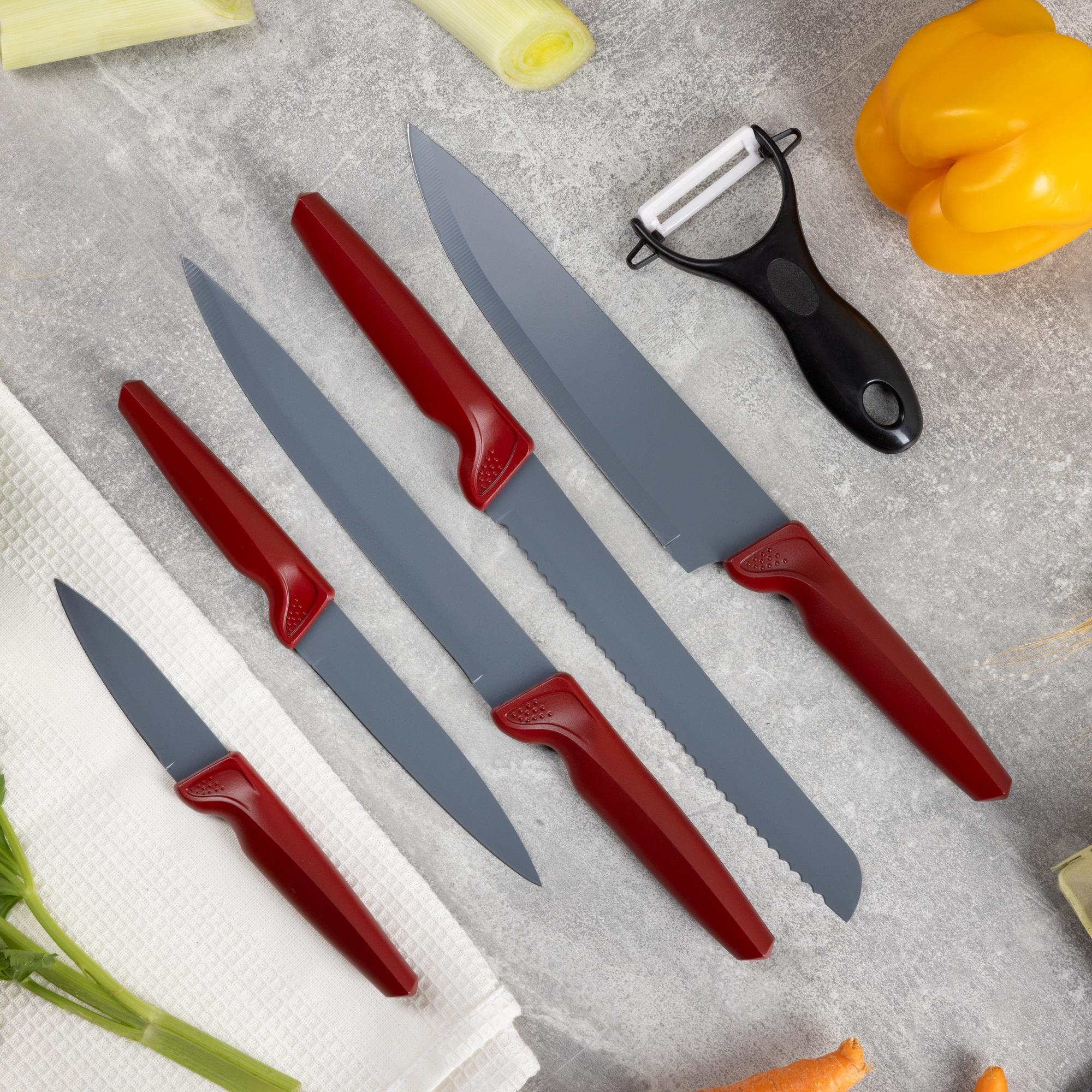 Juego de cuchillos de cocina con mangos rojos, un pelador y verduras como puerro, pimiento amarillo y zanahorias sobre una superficie gris.