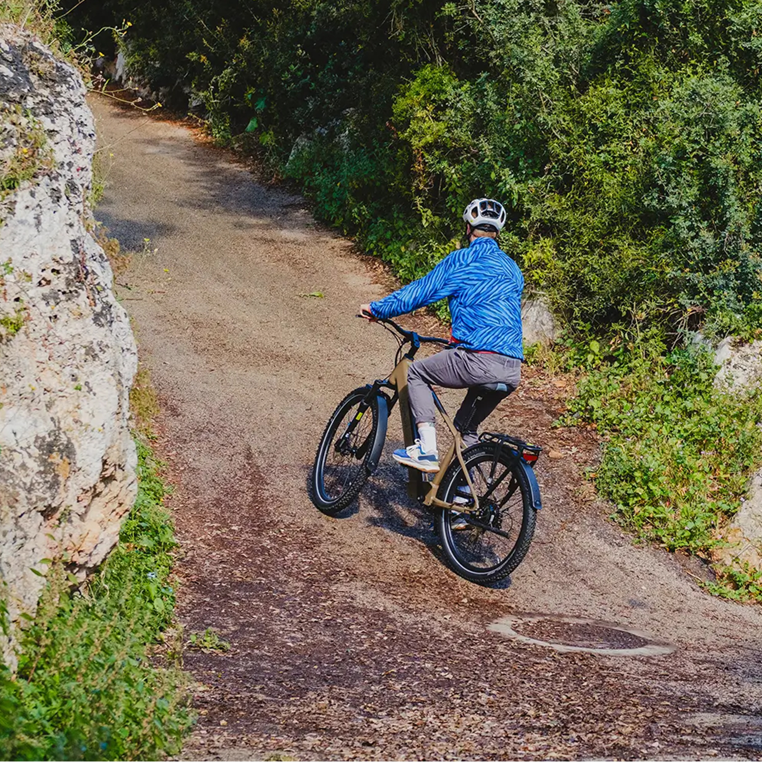 Eine Person fährt mit dem Fahrrad auf einem Feldweg. Der Weg ist von einer Felswand und dichtem Grün gesäumt. Der Radfahrer trägt einen Helm.