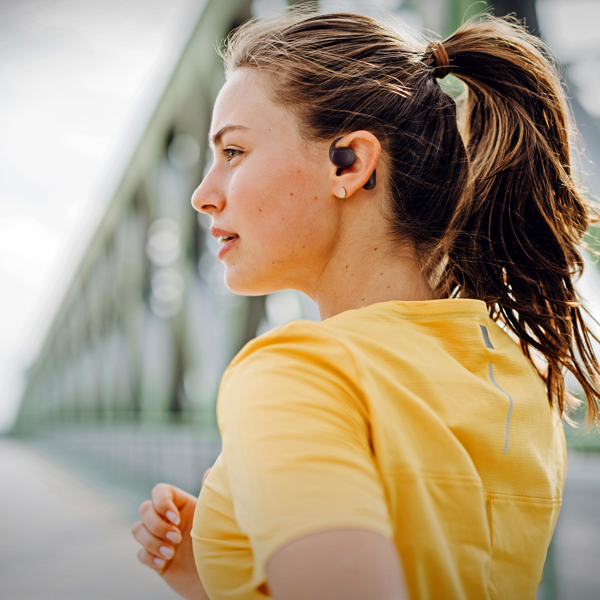 Una mujer con coleta, camisa amarilla y auriculares corre al aire libre. Un puente está al fondo.