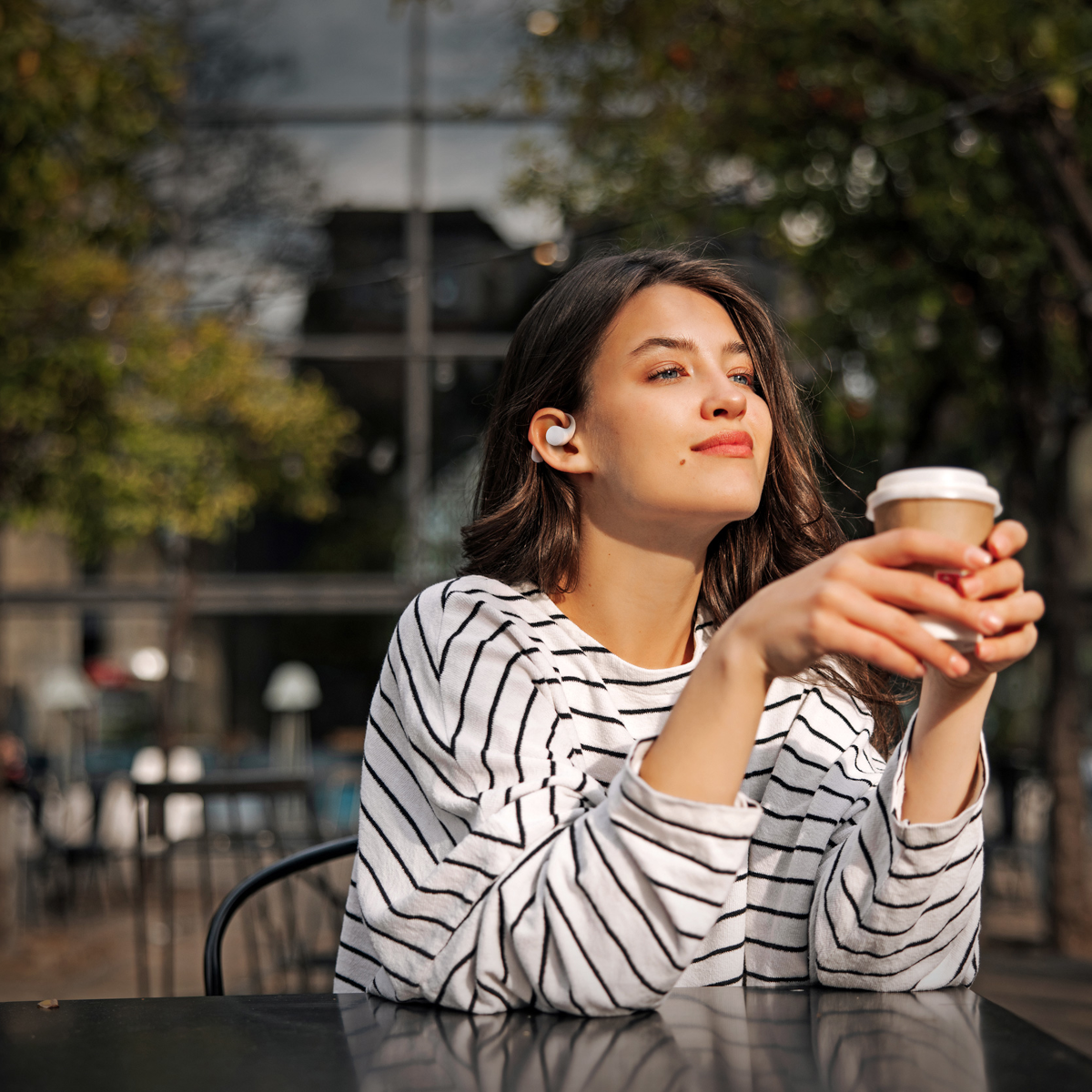 Una mujer con auriculares, camisa a rayas y taza de café está sentada al aire libre, mirando hacia arriba. Árboles y un edificio al fondo.