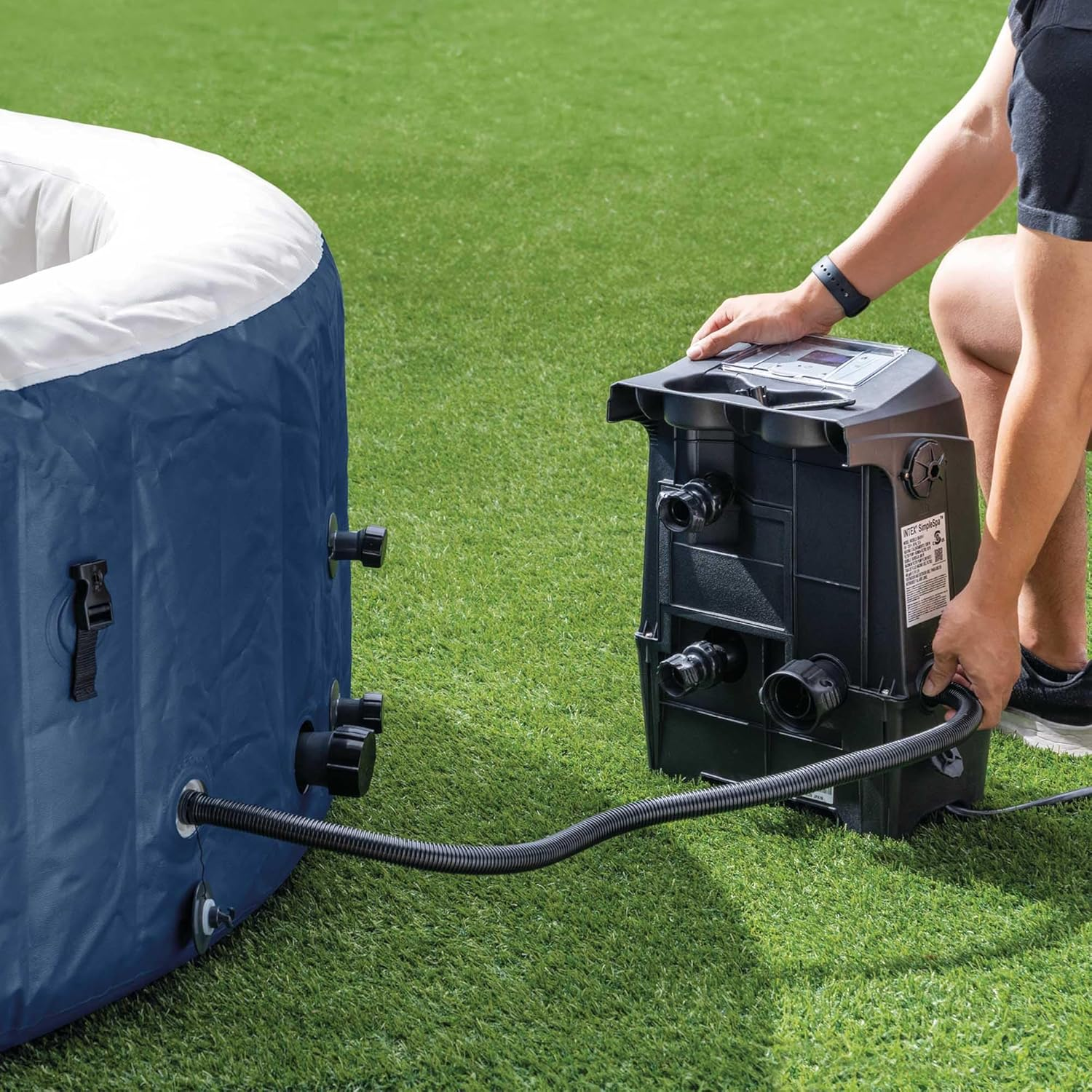 Eine Person verbindet eine schwarze Pumpe mit einem blauen aufblasbaren Whirlpool auf grünem Gras. Die Pumpe hat ein Digitaldisplay.