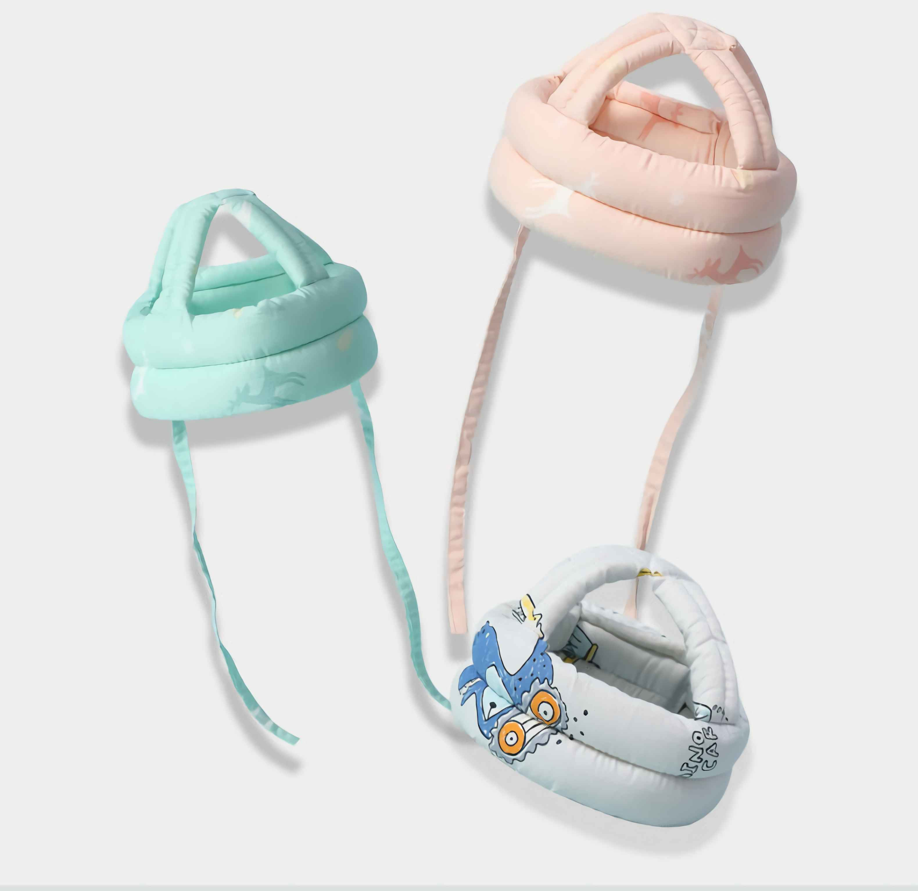Drei Babyhelme: Minze, Rosa und Weiß. Die Helme haben Riemen und sind mit Mustern und Illustrationen verziert.