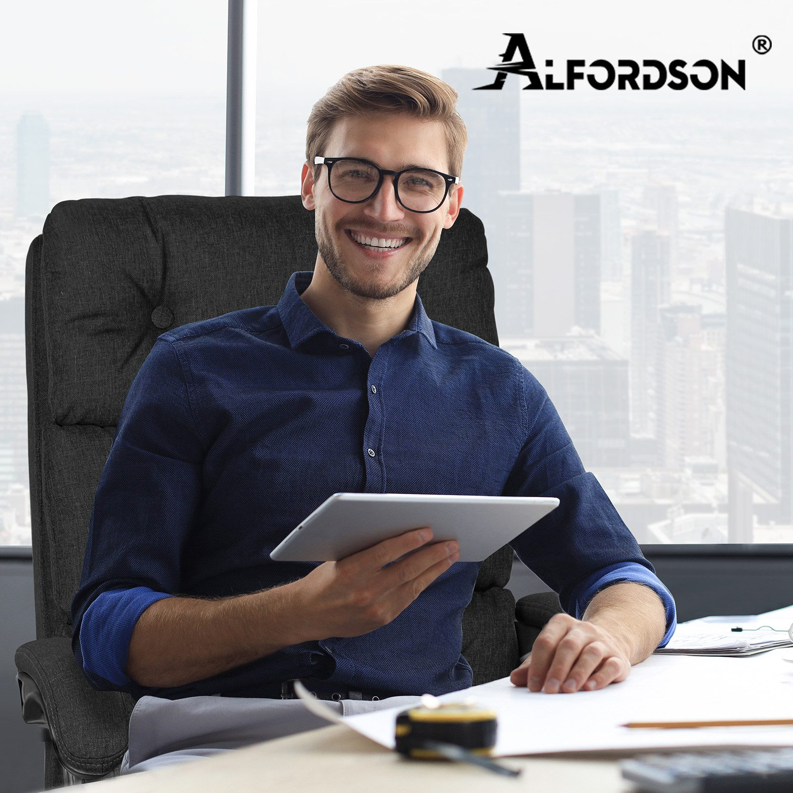 Ein Mann lächelt und hält ein Tablet. Er trägt eine Brille und ein blaues Hemd und sitzt in einem Bürostuhl mit Stadtblick.