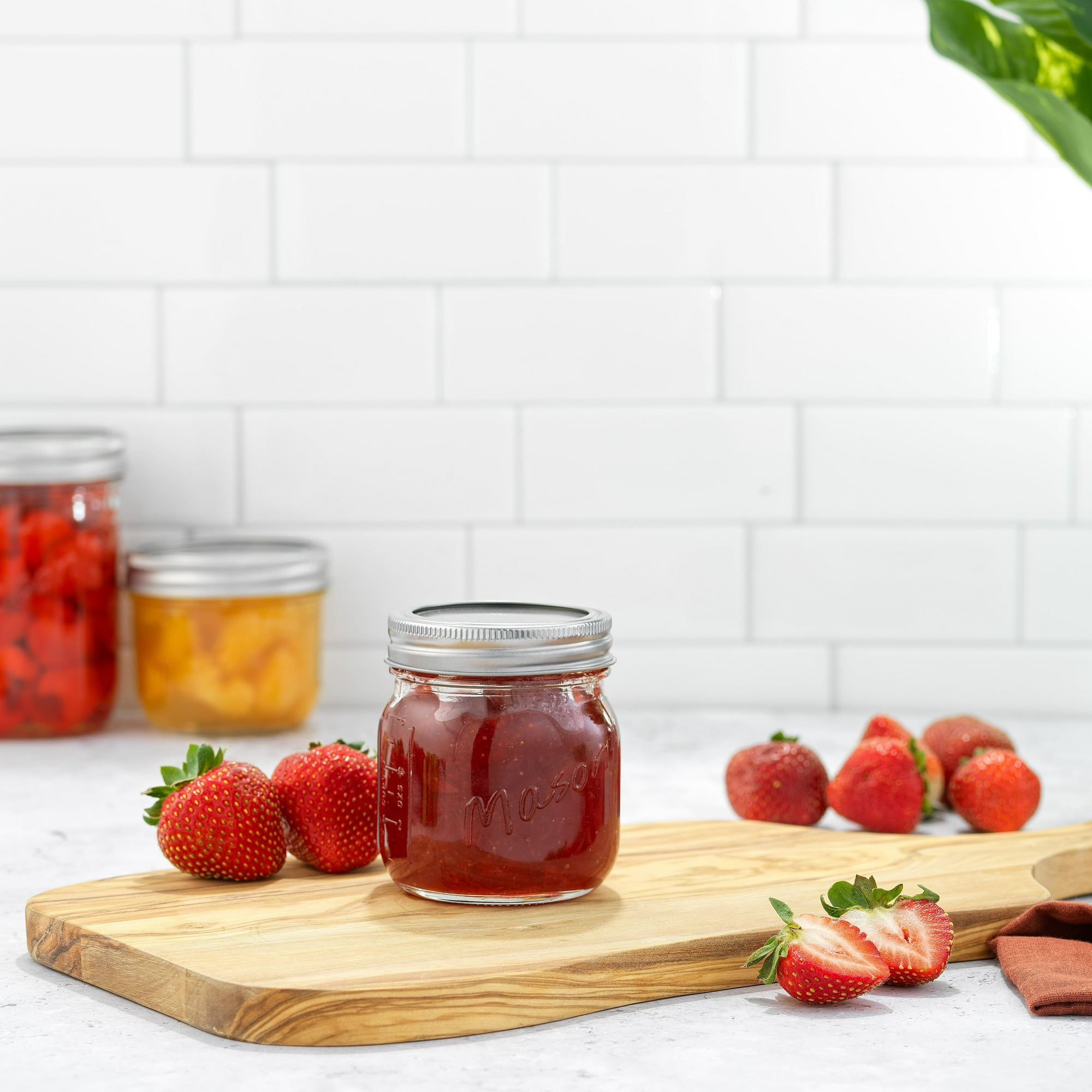 Ein Glas Erdbeermarmelade auf einem Holzbrett mit Erdbeeren. Andere Gläser im Hintergrund. Weißer Ziegelwandhintergrund.