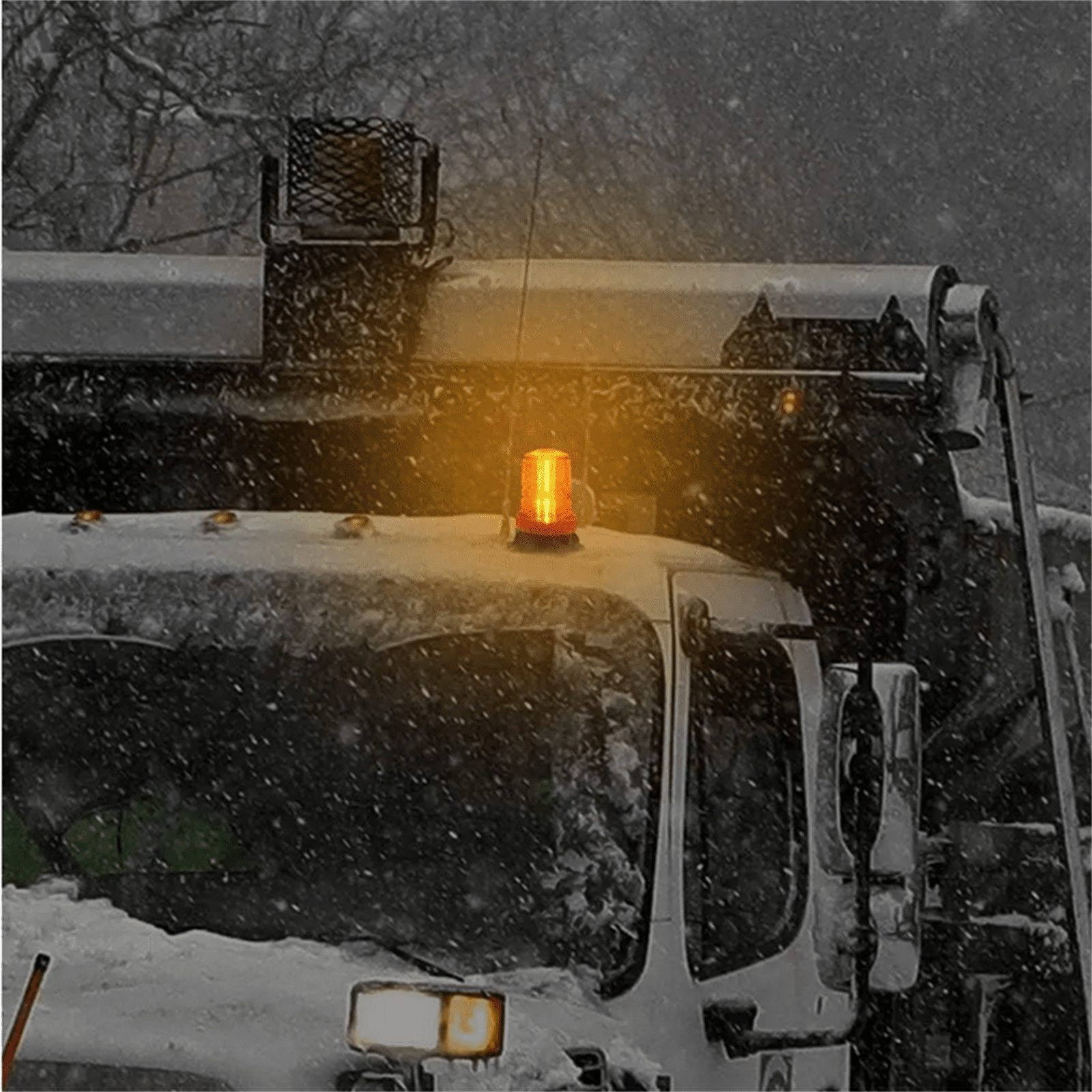 Ein Schneepflug mit orangefarbenem Blinklicht, in einer verschneiten Umgebung. Schnee fällt.