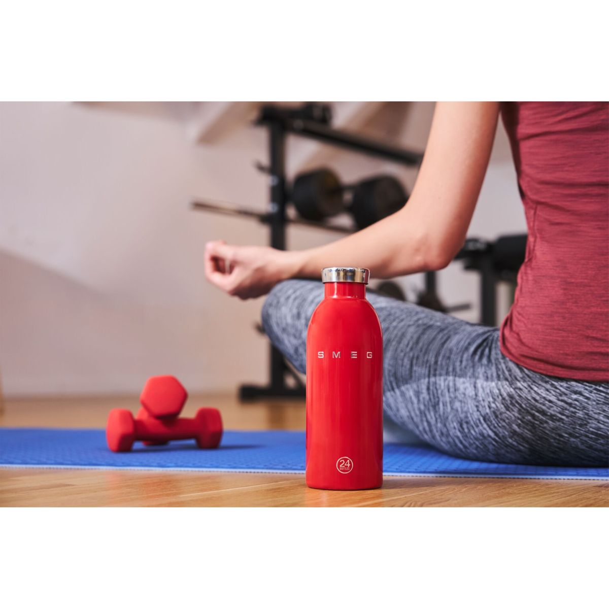 Eine Person sitzt mit einer roten Wasserflasche, einer Hantel und einer Yogamatte in einem Raum. Die Person trägt ein rotes Hemd und graue Leggings.