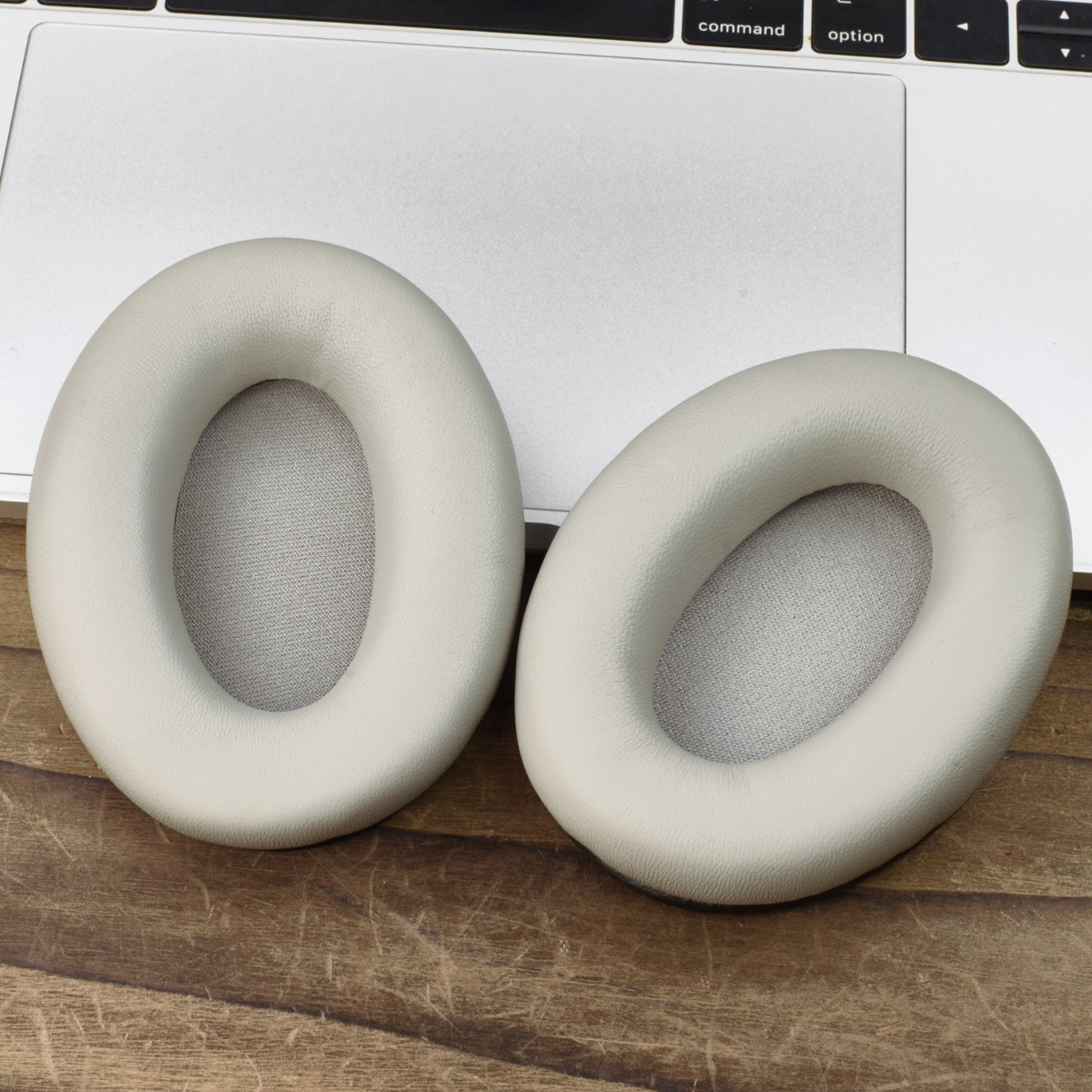 Dos almohadillas beige para auriculares sobre el teclado de un portátil. Las almohadillas son ovaladas, con tela gris en el interior. Suelo de madera en primer plano.