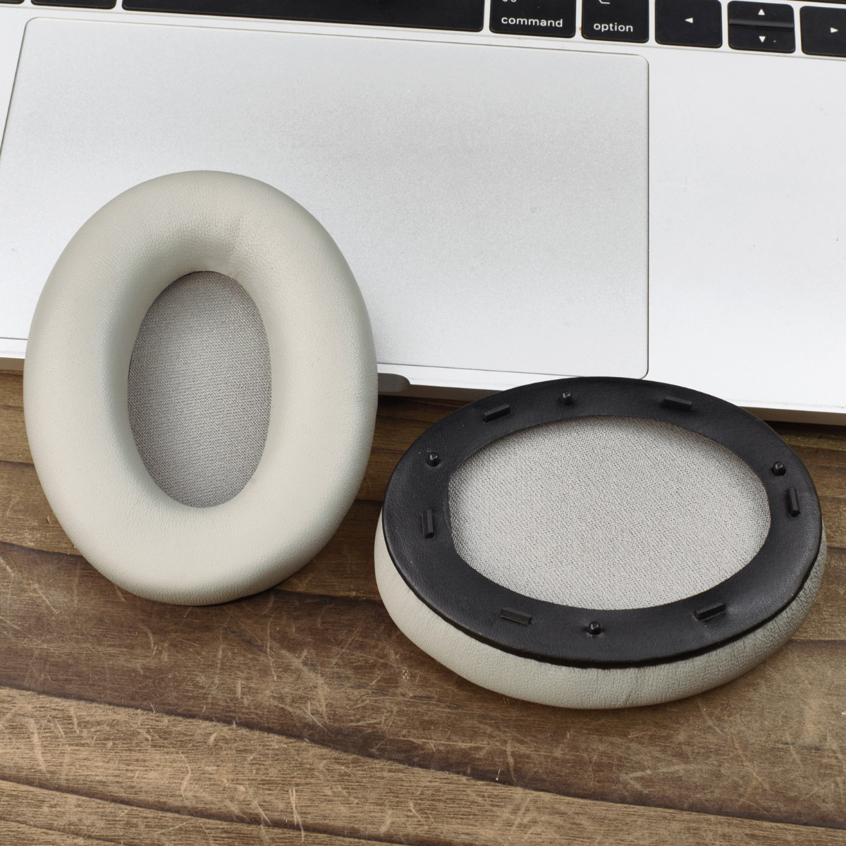 Dos almohadillas beige para auriculares están sobre una superficie de madera junto a una laptop. Una almohadilla está desmontada.