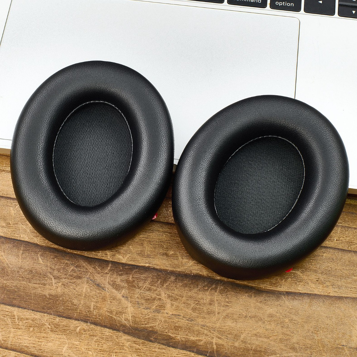 Dos almohadillas negras para auriculares sobre una superficie de madera, junto a un teclado de portátil. Las almohadillas son ovaladas.