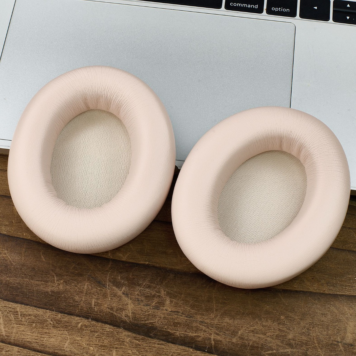Dos almohadillas rosas para auriculares están sobre una mesa de madera, junto a una computadora portátil plateada. Las almohadillas son ovales con interior de malla.