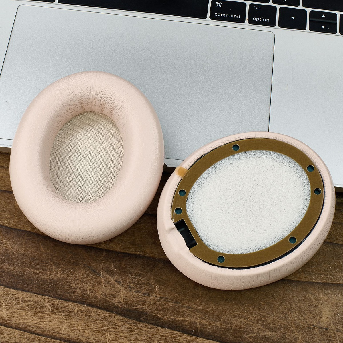 Dos almohadillas rosas para auriculares, una desmontada. Descansan sobre una superficie de madera junto a una laptop.
