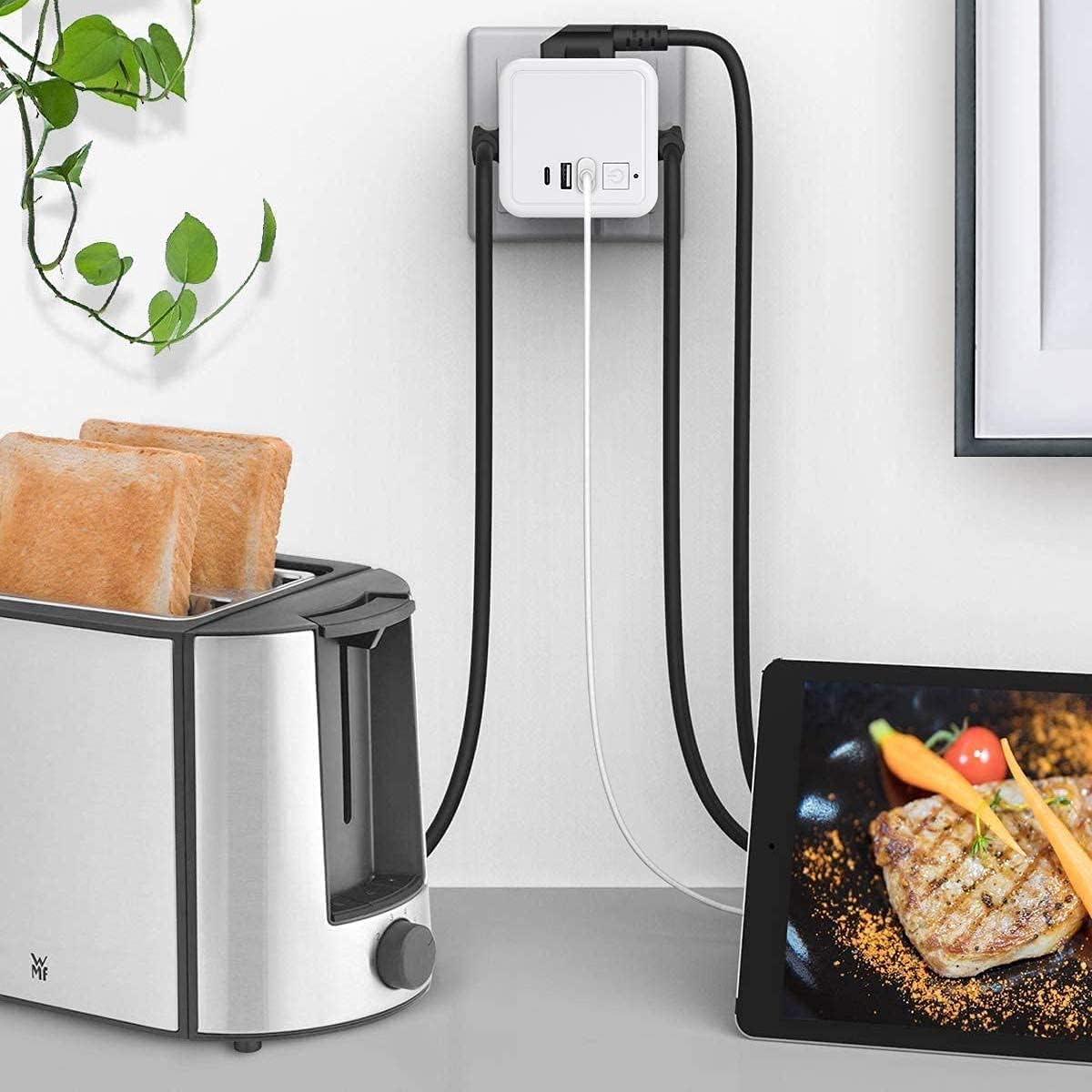 Toaster mit Brot, Steckdose mit Kabeln, Tablet mit Essen auf einem Tisch. Grüne Pflanze an der Wand. Ein Bilderrahmen rechts.