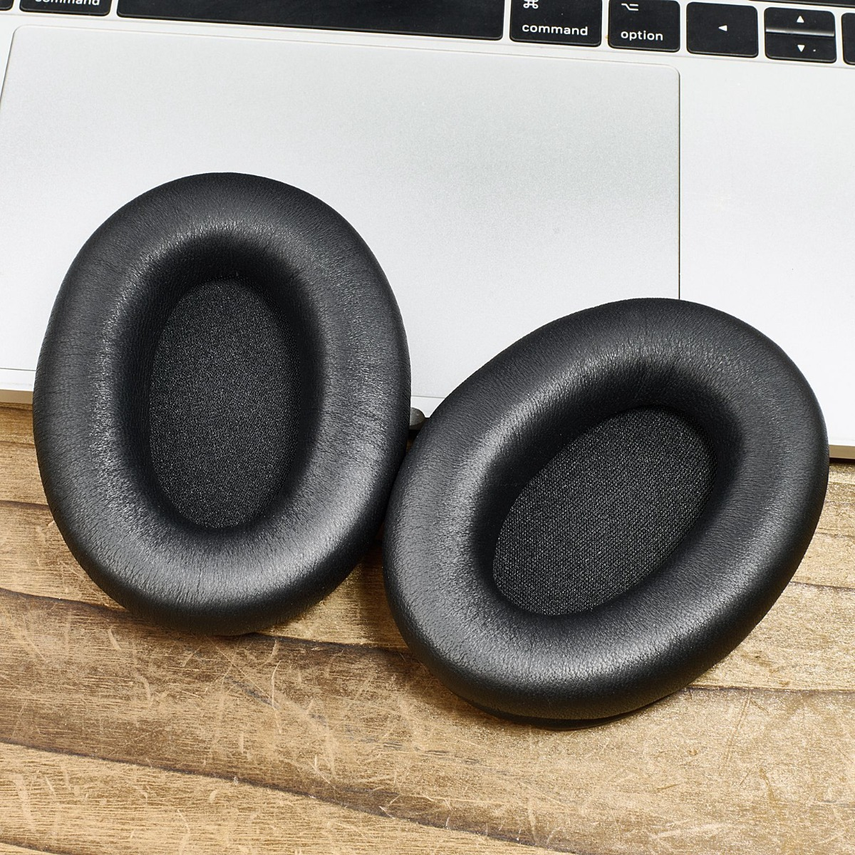 Zwei schwarze Kopfhörer-Ohrmuscheln auf einer Holzoberfläche neben einer Laptop-Tastatur. Die Ohrmuscheln sind oval und lederartig.