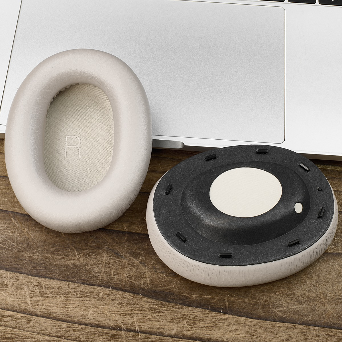 Zwei beige Kopfhörer-Ohrmuscheln und ein schwarz-beiges Teil des Kopfhörers. Im Hintergrund ist ein Laptop. Alles liegt auf einem Holztisch.