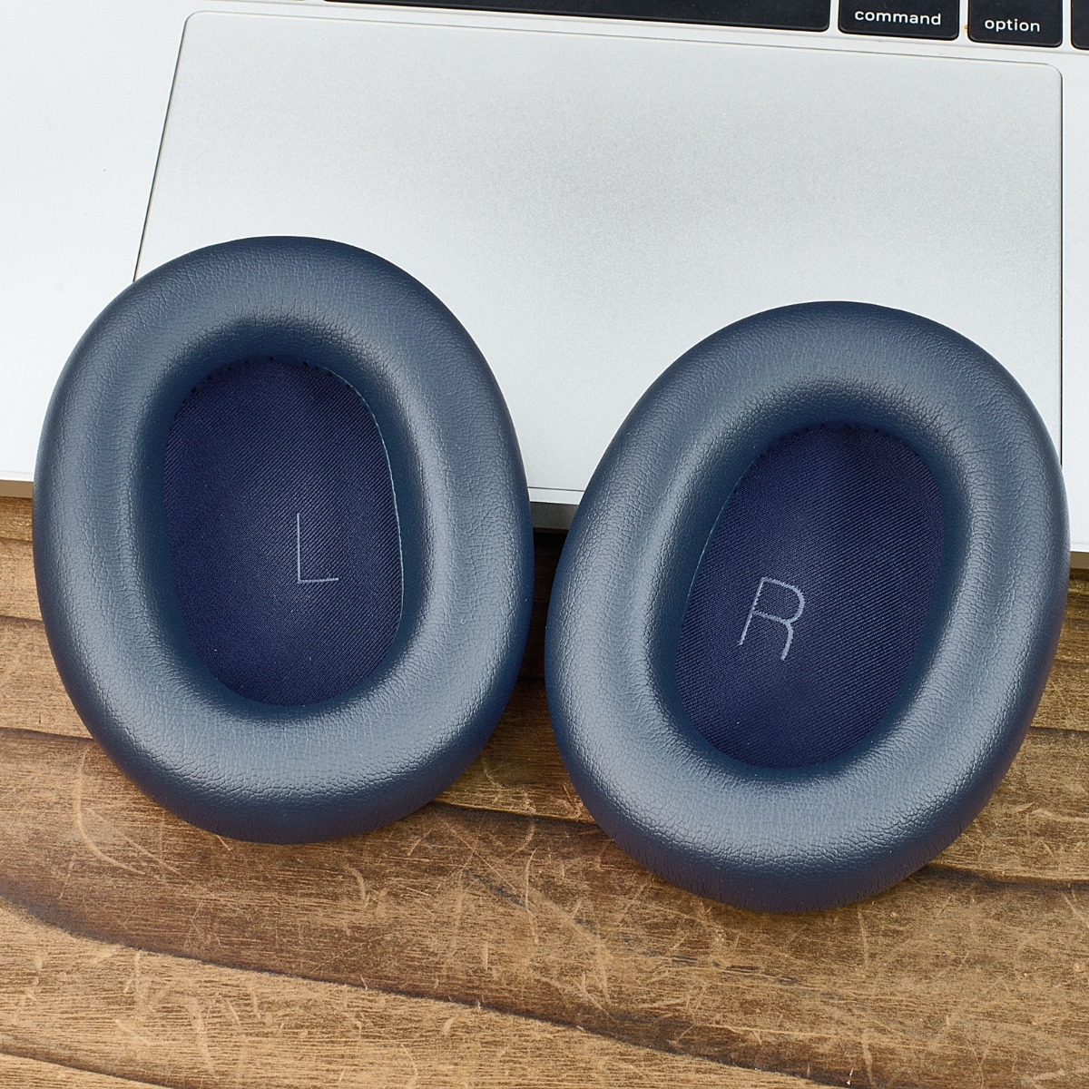 Zwei blaue Kopfhörer-Ohrmuscheln auf einer Holzoberfläche, im Hintergrund eine Laptop-Tastatur. Die linke Ohrmuschel hat ein 'L', die rechte ein 'R'.