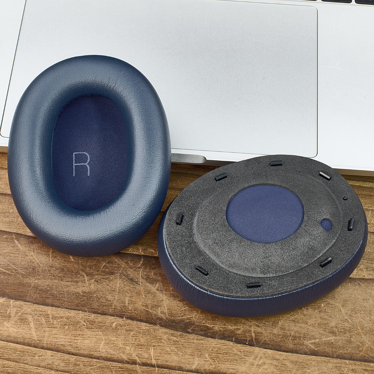 Zwei blaue Kopfhörer-Ohrmuscheln auf einer Holzoberfläche neben einem Laptop. Eines hat ein 'R', das andere schwarzen Schaumstoff.