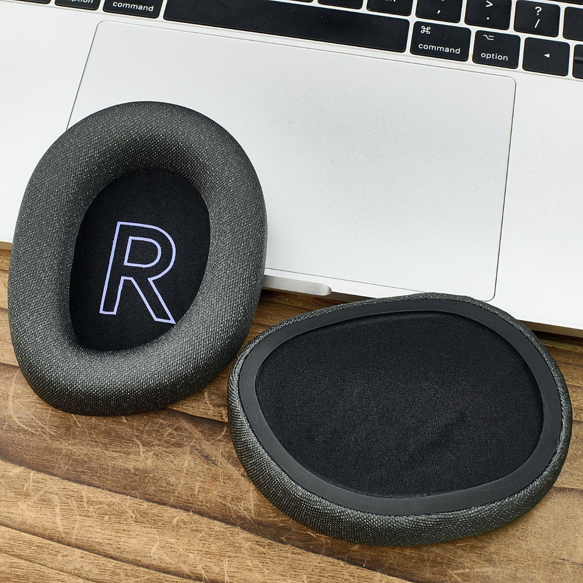 Dos almohadillas grises para auriculares sobre madera y un portátil. Una tiene una 'R' blanca sobre tela negra.