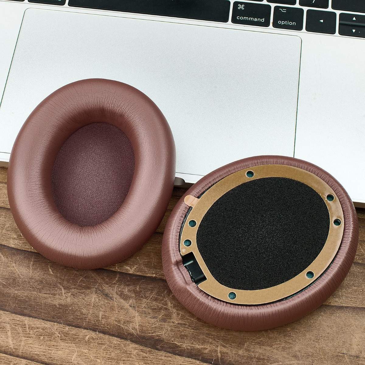 Dos almohadillas marrones para auriculares sobre una superficie de madera junto a un teclado de portátil. Una almohadilla está suelta.