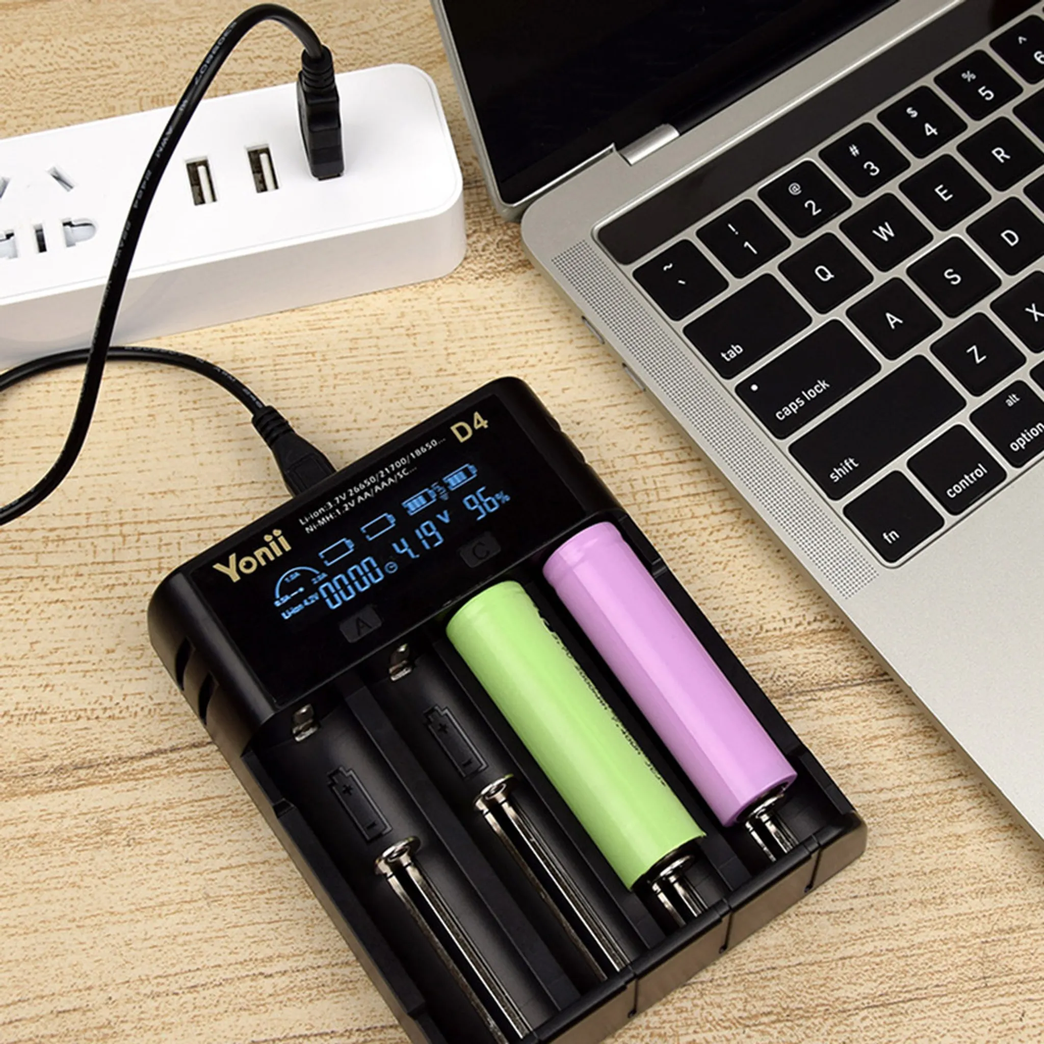 Ein Batterieladegerät mit zwei Batterien, ein Laptop und eine Steckdose befinden sich auf einer Holzoberfläche.