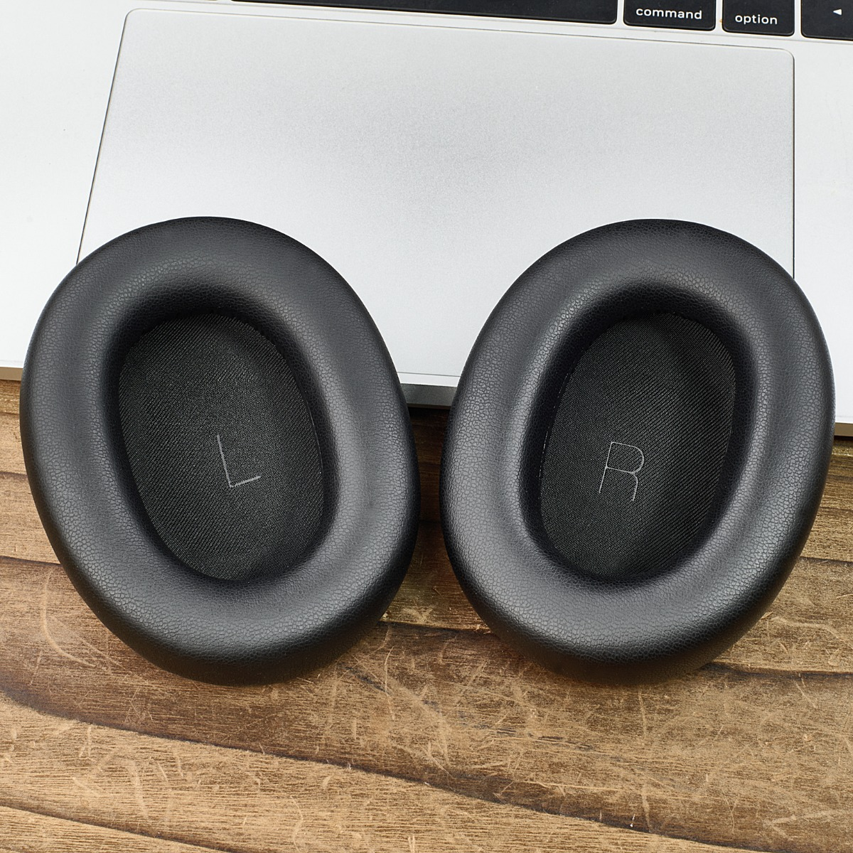 Dos almohadillas negras para auriculares con marcas 'L' y 'R', sobre un teclado de portátil y superficie de madera.