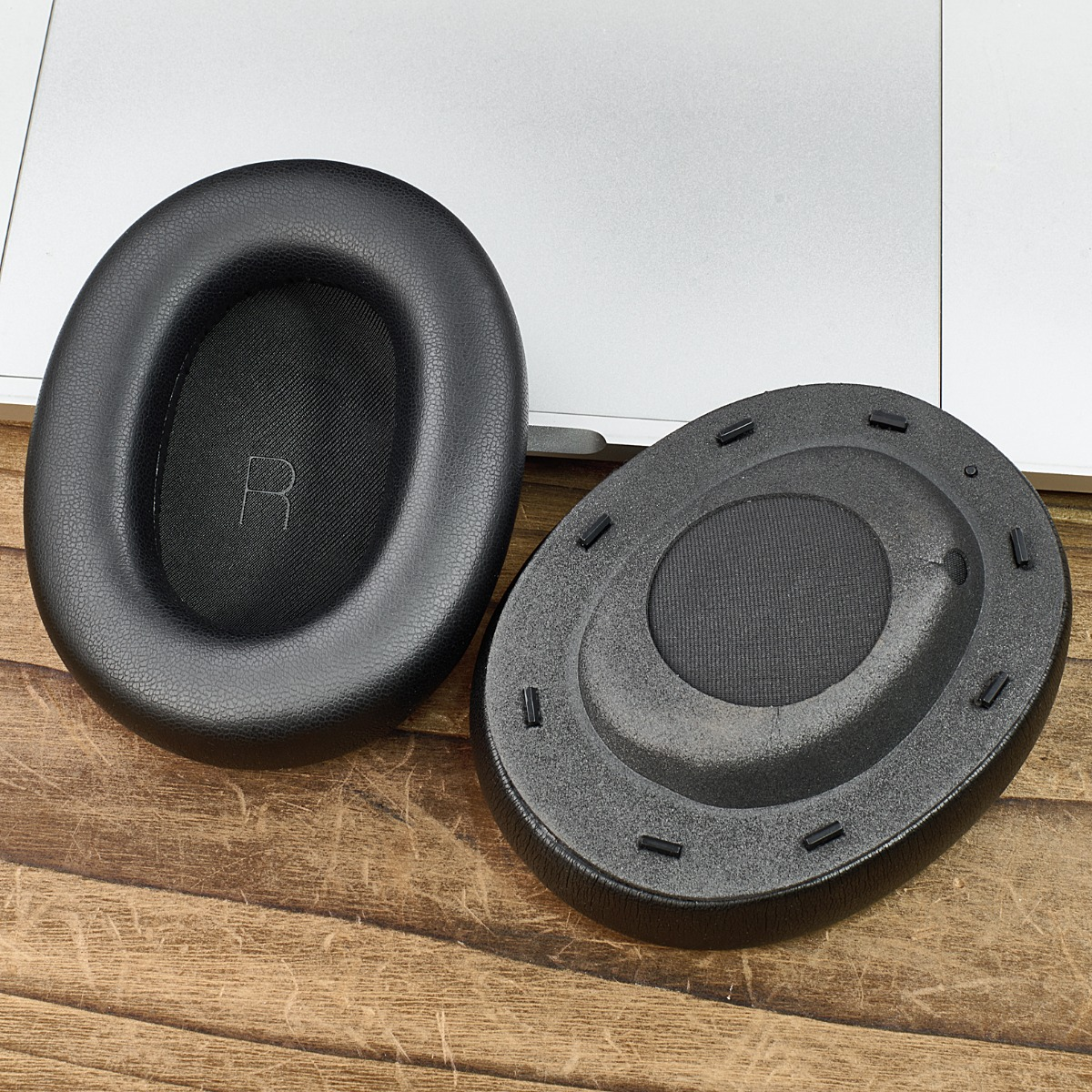 Dos almohadillas negras para auriculares sobre una superficie de madera, una con una 'R' y otra con pequeños pasadores, cerca de una laptop plateada.