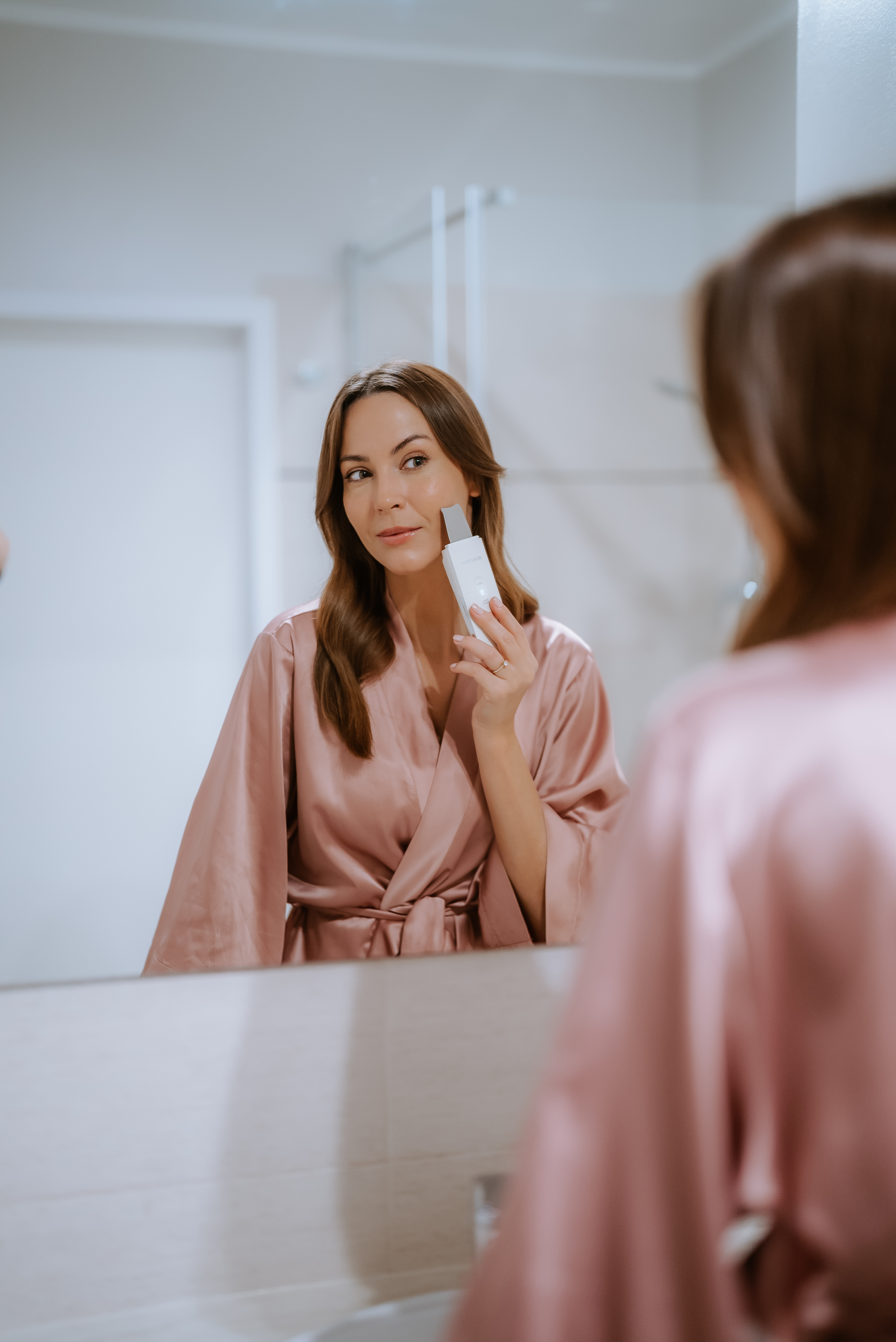 Mujer con bata rosa usando un dispositivo de limpieza facial frente a un espejo en el baño.