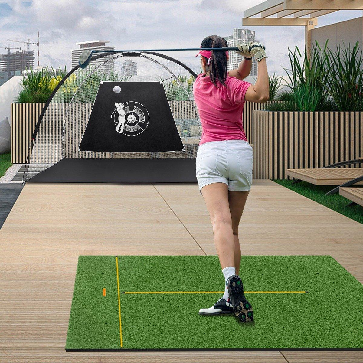 Una mujer con camisa rosa y pantalones cortos blancos golpea una pelota de golf sobre una alfombra verde. Una red y un blanco detrás de ella. Edificios al fondo.