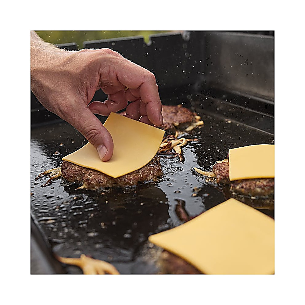Una mano posiziona una fetta di formaggio giallo su un hamburger su una piastra. Sono visibili altri hamburger e fette di formaggio.