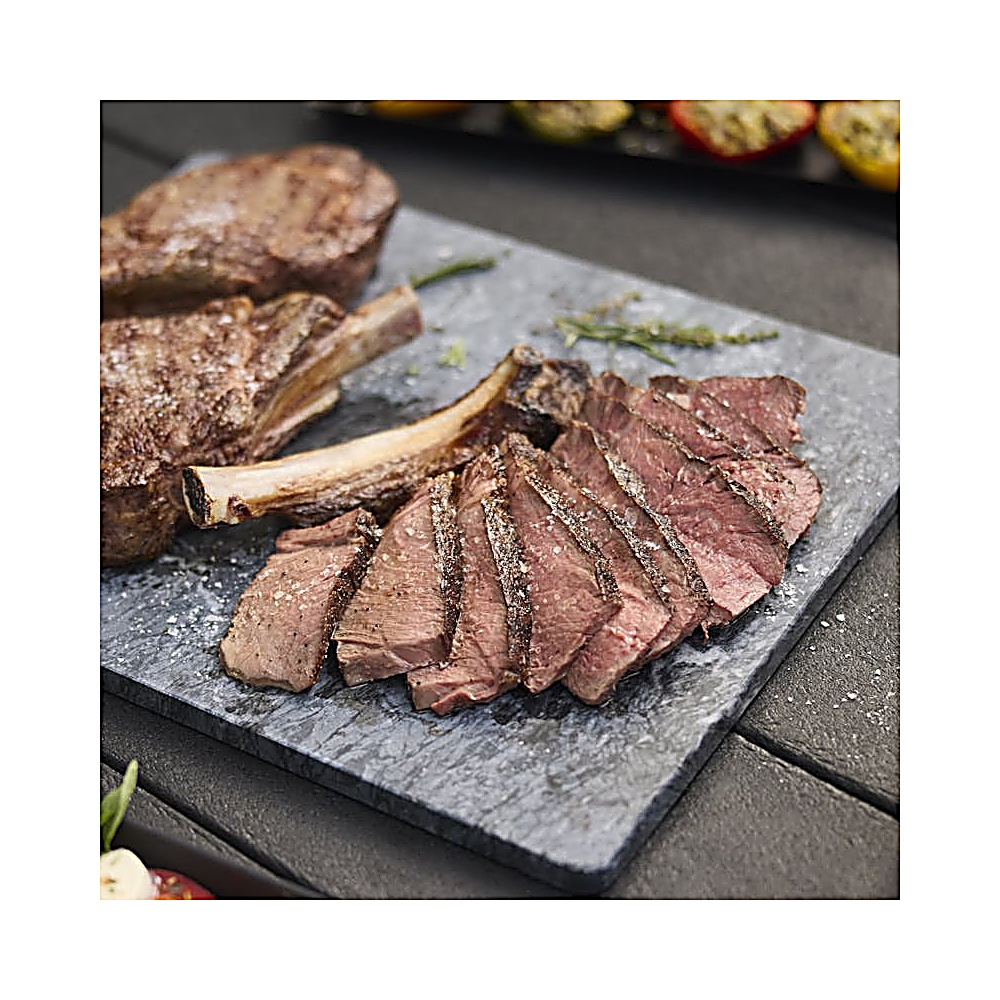 Bistecca alla griglia a fette su una tavola di pietra grigia. Altre bistecche e verdure grigliate sullo sfondo. Sale cosparso sulla carne.