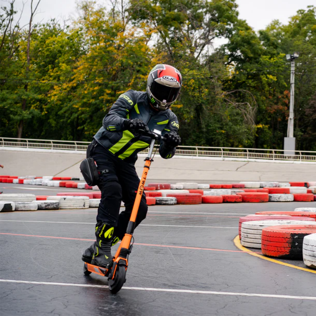 Osoba w kasku jedzie na pomarańczowej hulajnodze po torze. Tor ma bariery z opon. W tle drzewa.