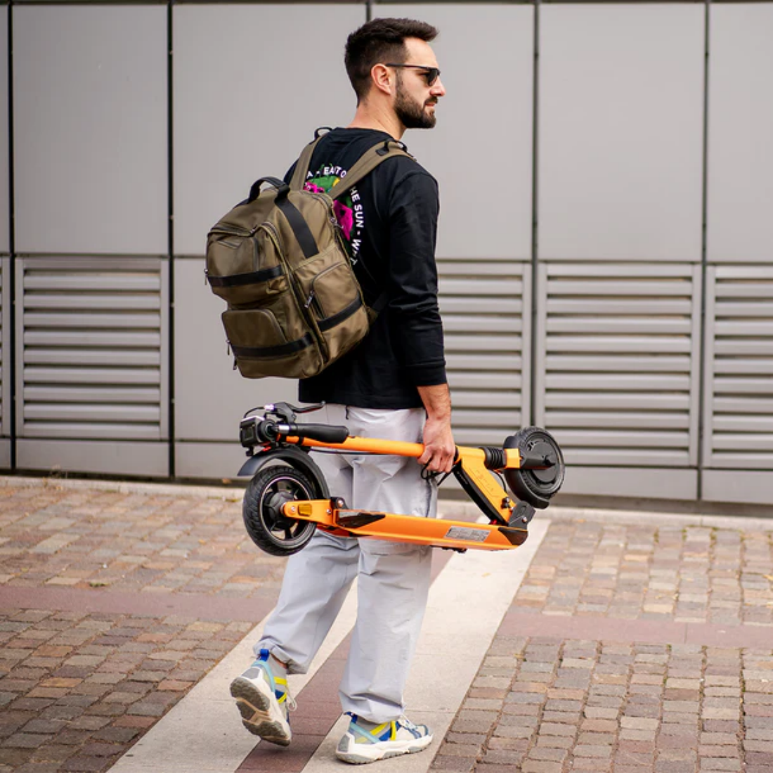 Mężczyzna z plecakiem niesie pomarańczową hulajnogę elektryczną. Ma okulary przeciwsłoneczne, czarną koszulę, jasne spodnie i trampki.