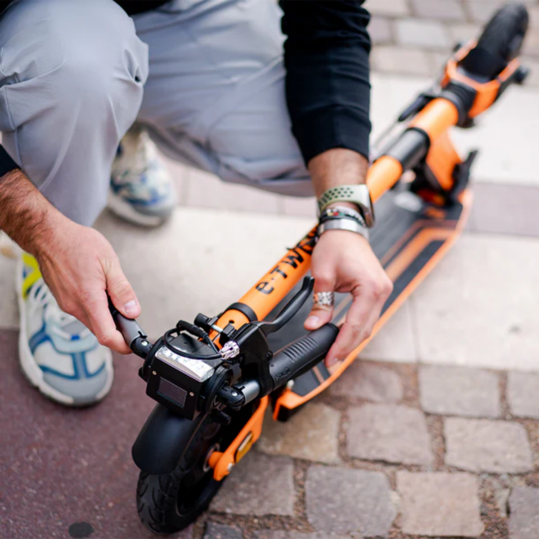 Osoba reguluje pomarańczowo-czarną hulajnogę elektryczną. Osoba ma na sobie szare spodnie i trzyma kierownicę na ceglanej powierzchni.