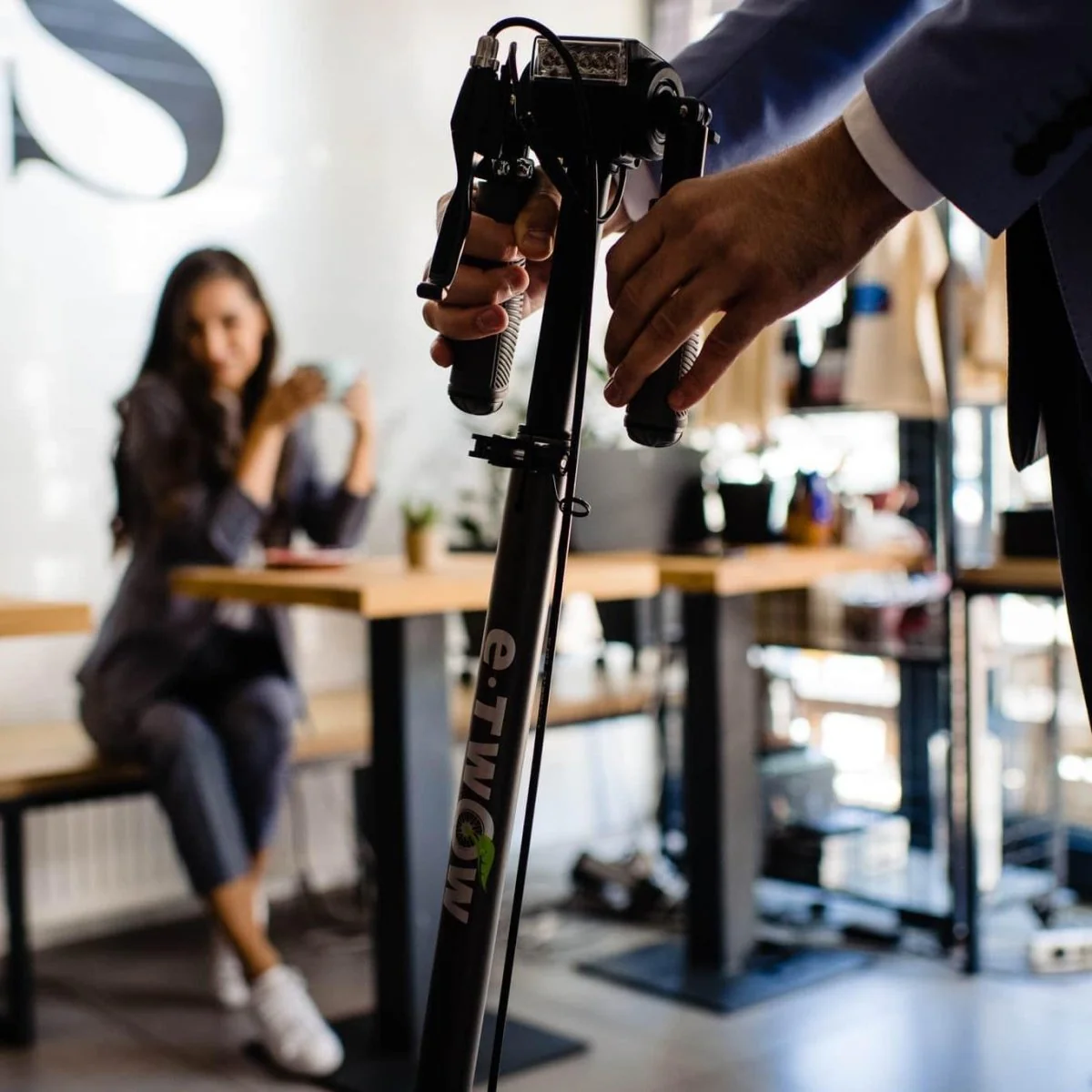 Osoba trzyma czarną hulajnogę elektryczną. Kobieta siedzi przy stole w tle, trzymając filiżankę.