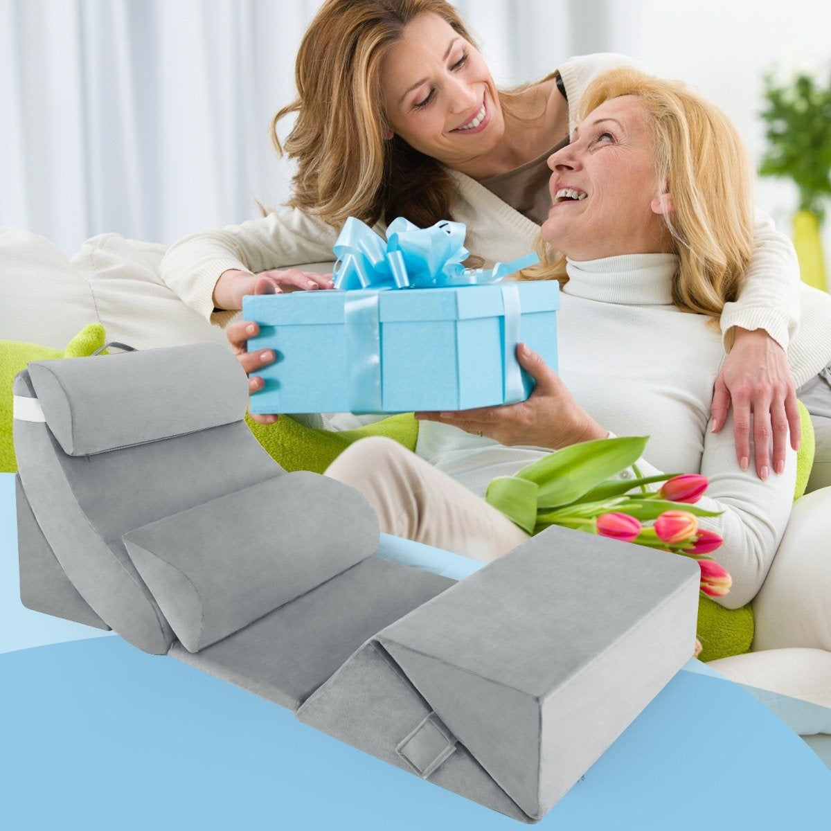 Una mujer le da una caja de regalo azul claro a otra mujer. Una cuña de cama gris y flores están en primer plano. Ambas mujeres sonríen.