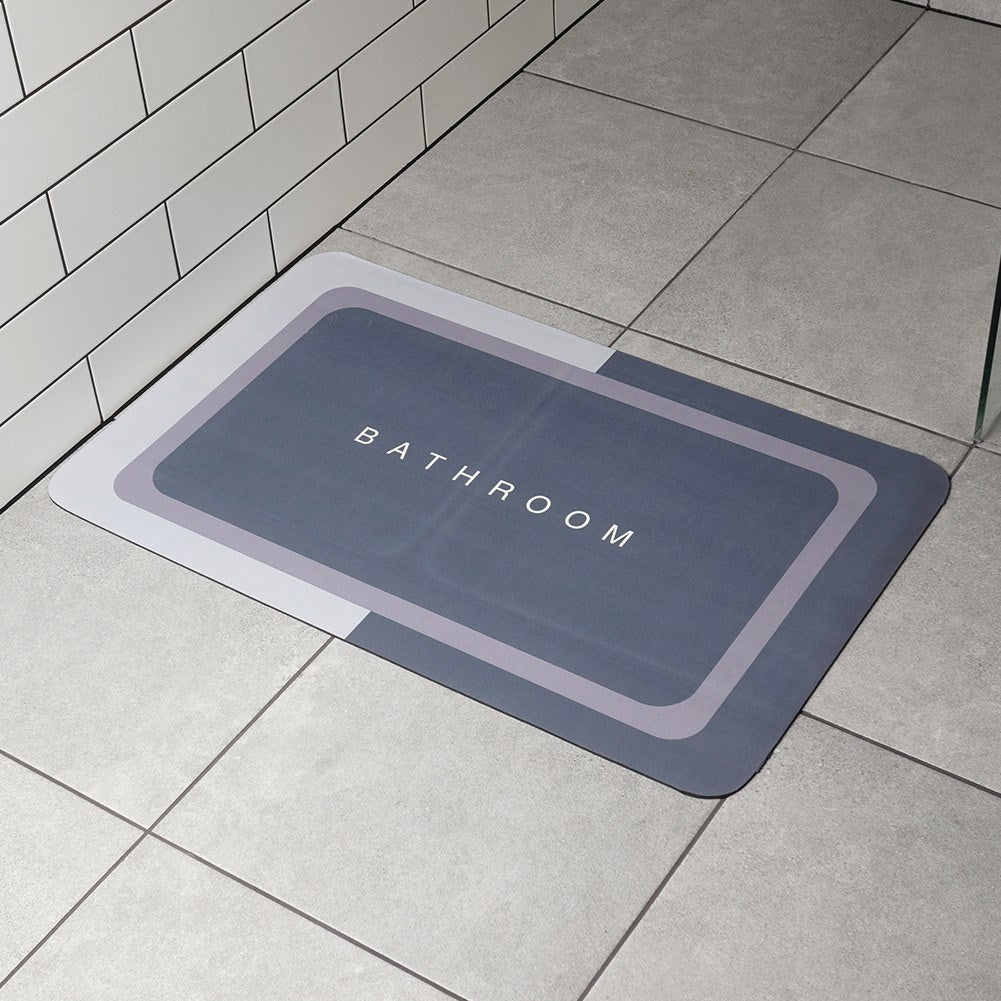 Una alfombra de baño gris con 'BATHROOM' en letras blancas sobre baldosas grises. Una pared de azulejos blancos a la izquierda.